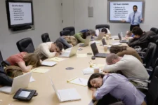 Meeting attendees asleep at a conference table while a presenter shows a text-heavy bullet-point slide